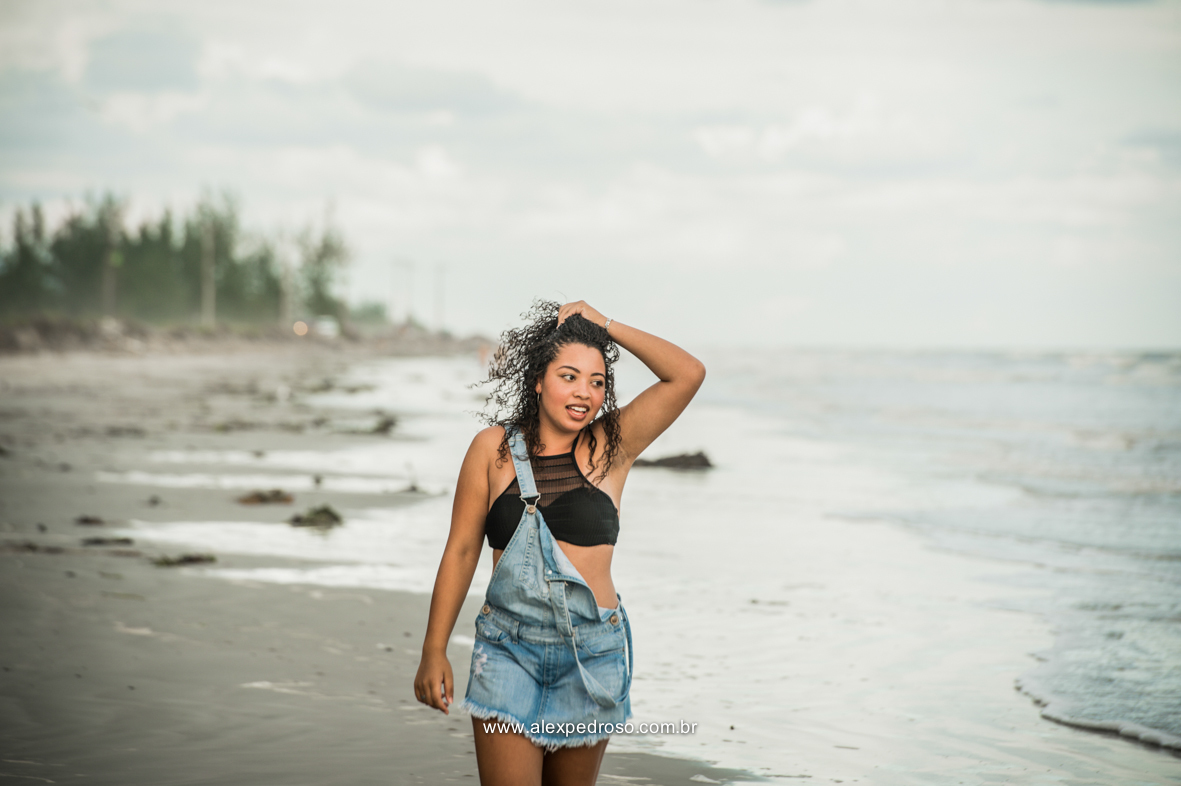 Menina de cabelo cacheado mexendo em seu próprio cabelo, andando pela praia, usando um cropped preto e um macacão jeans, olhando para o mar, foto tirada do joelho pra cima.