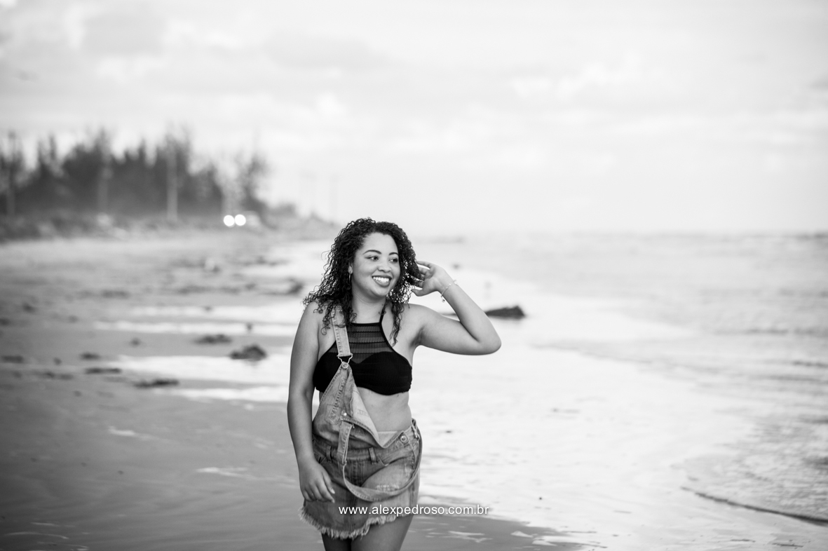 Menina de cabelo cacheado mexendo em seu próprio cabelo, andando pela praia, usando um cropped preto e um macacão jeans, olhando para o mar, foto tirada do joelho pra cima e em preto e branco