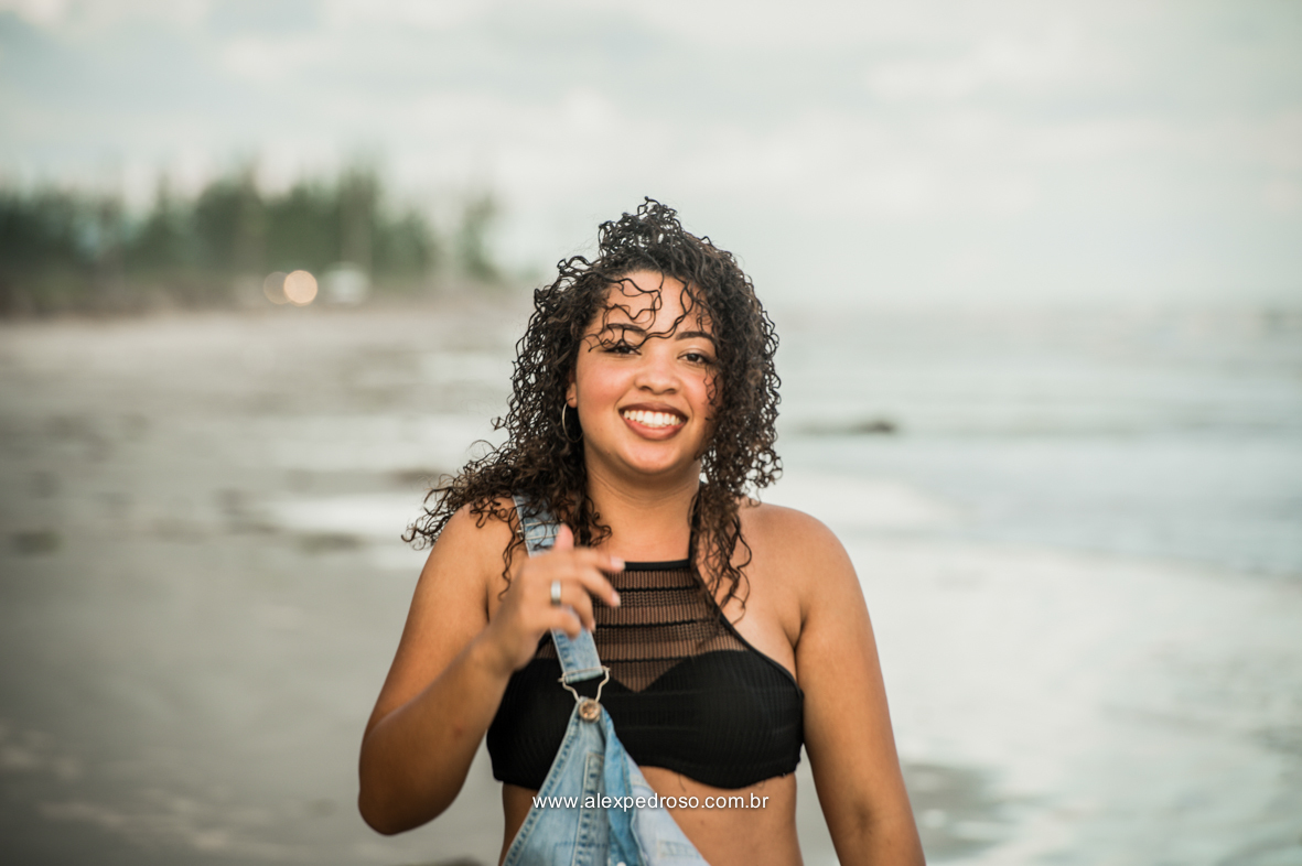 Menina de cabelo cacheado sorrindo, no meio de uma praia usando um cropped preto e um macacão jeans, foto tirada da barriga pra cima.