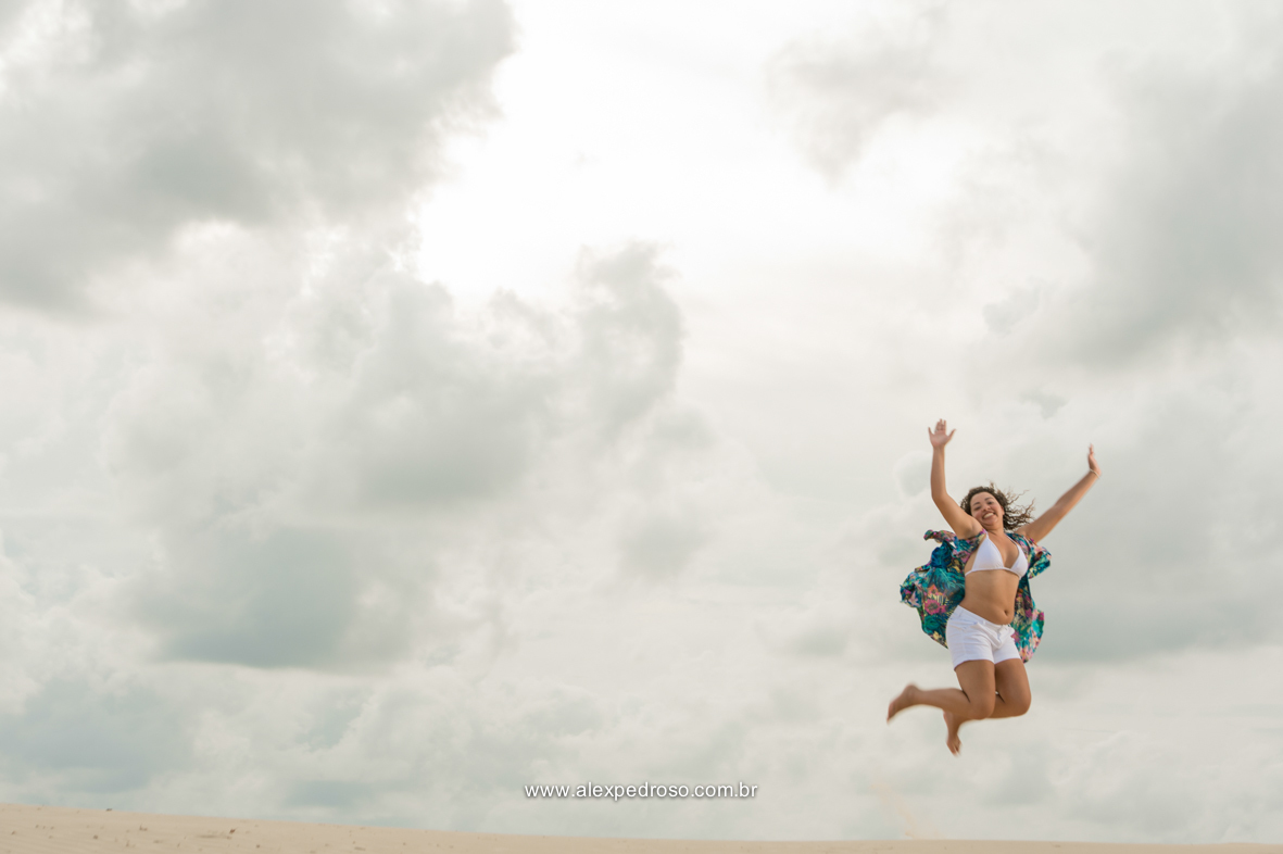Menina de cabelo cacheado pulando na areia, usando a parte de um biquíni branca e um short branco, com uma saída de praia verde, foto tirada em praia.