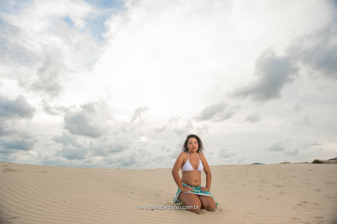 Menina de cabelo cacheado sentada de joelhos na areia com as mãos na perna, usando a parte de um biquíni branco e uma saia verde, olhando pra paisagem e com a cara séria, foto tirada em uma praia.