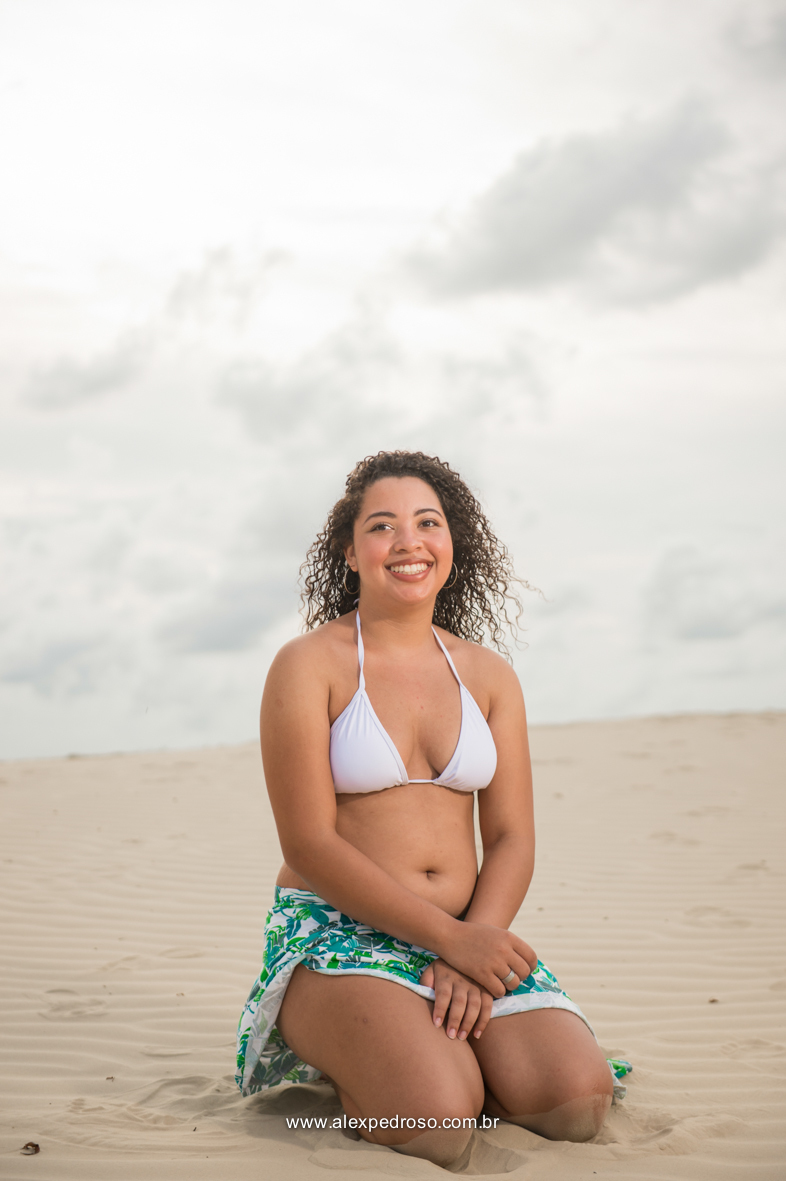 Menina de cabelo cacheado sorrindo com o biquíni branco, foto tirada do umbigo pra cima e realizada em uma praia.