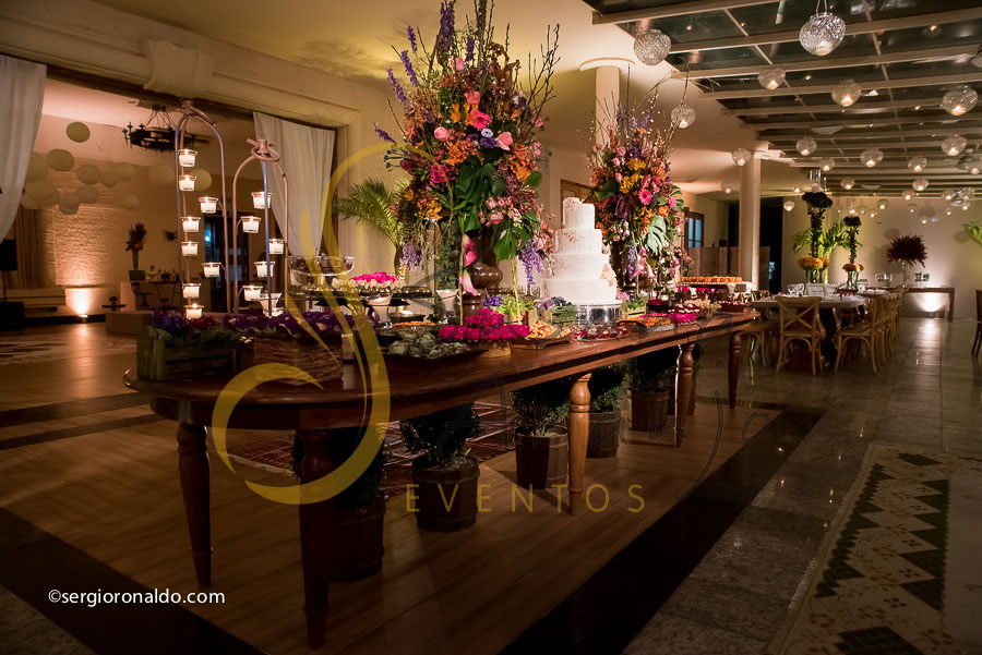 mesa de doces e bolo com flores e arranjos de flores rosa, laranja, lilás. roxo, e mobiliário de madeira. 