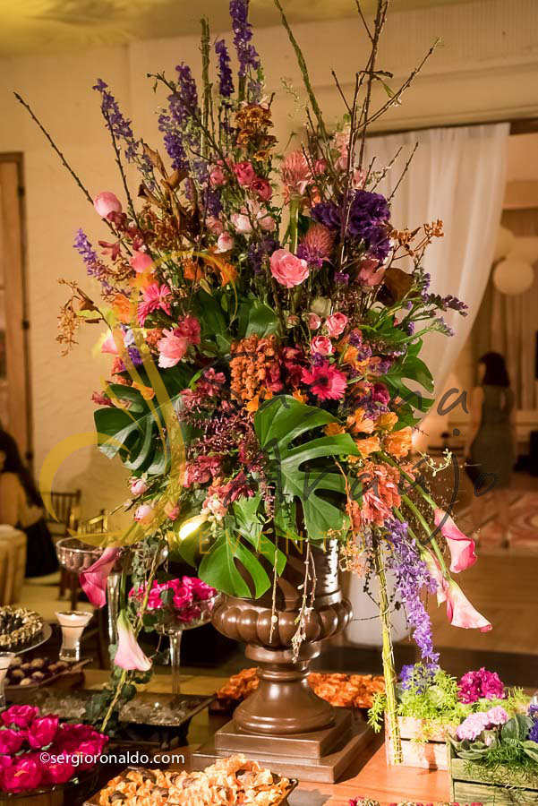 mesa de doces e bolo com flores e arranjos de flores rosa, laranja, lilás. roxo, e mobiliário de madeira. 