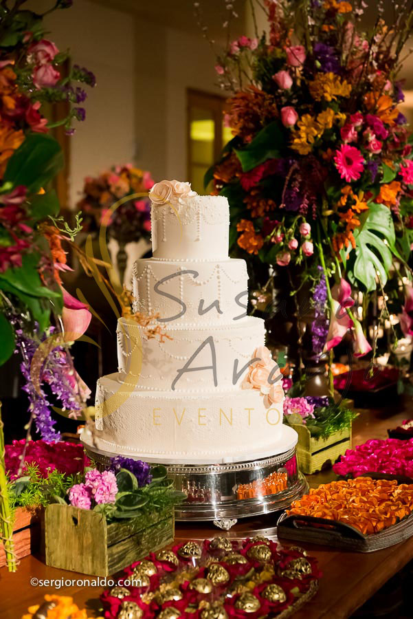 mesa de doces e bolo com flores e arranjos de flores rosa, laranja, lilás. roxo, e mobiliário de madeira. 