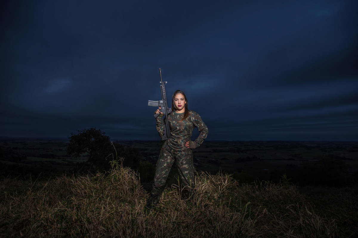 Sessão fotográfica no final de tarde em Piracicaba com céu azul escuro Brenda com uniforme camuflado segurando arma pra cima