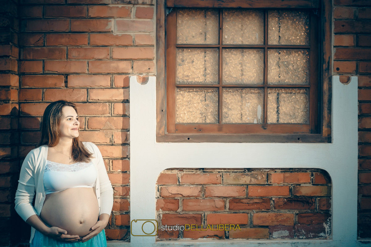 Cristiane segurando a barriga de gestante proximo a parede de tijolos, na sessão fotográfica feita em Piracicaba S.P.