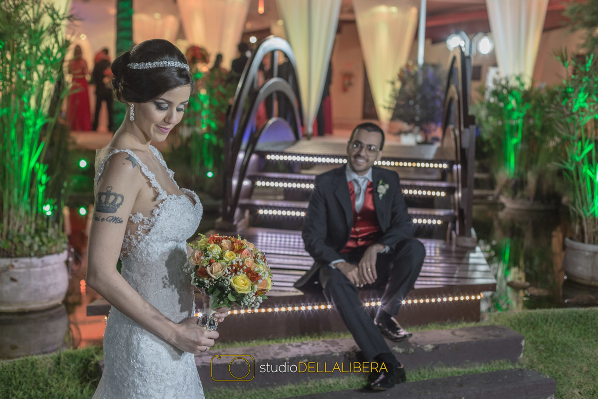 Noiva Camila segurando o Buque de flores coloridas na altura da cintura olhando para o buque com noivo sentado na ponte olhando para noiva com sorrizo no rosto na chácara Vó Dina em Nova Odessa SP, 