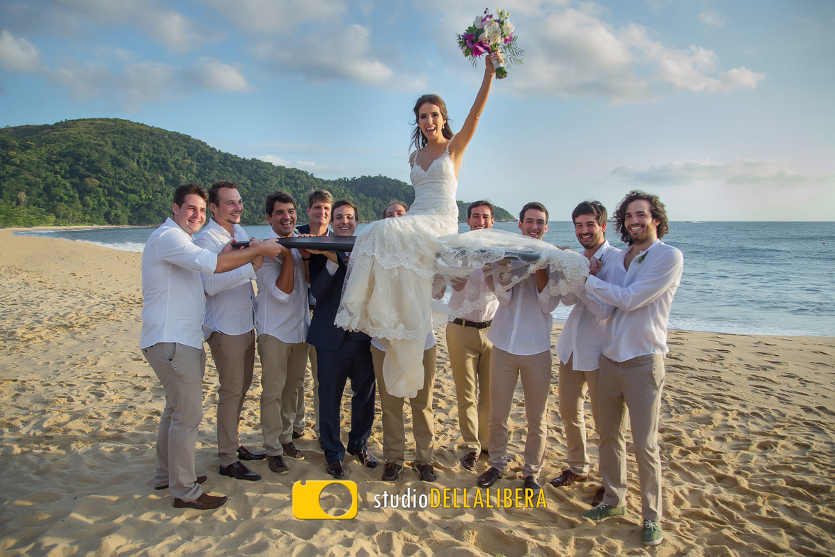 Noiva sendo carraga em cima de uma prancha de surf, com o noivo e os padrinhos levantndo a prancha na areia da praia em Maresias na reserva Tambá