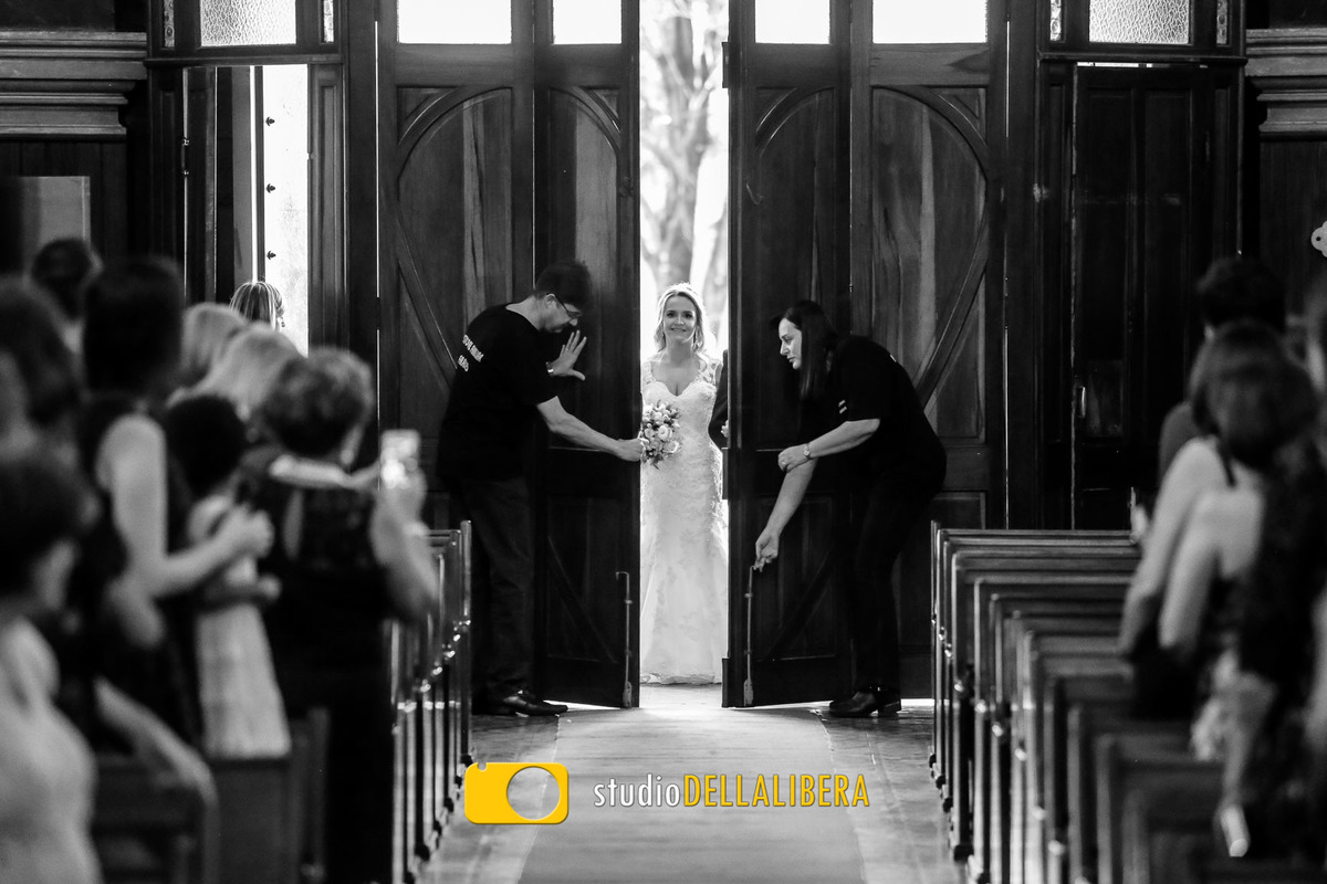 Porta da Igreja dos Frades de Piracicaba SP sendo aberta com a noiva aparecendo pela fresta, com os convidados em pé esperando a entrada da noiva na Igreja o noivo esperando no altar

