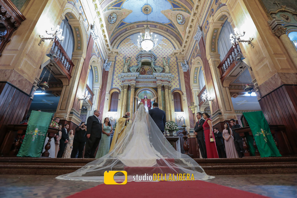 Igreja dos frades com a noiva de costas mostrando to o vestido da noiva com o véu esticado e o noivo com terno azul no altar e os padrinhos