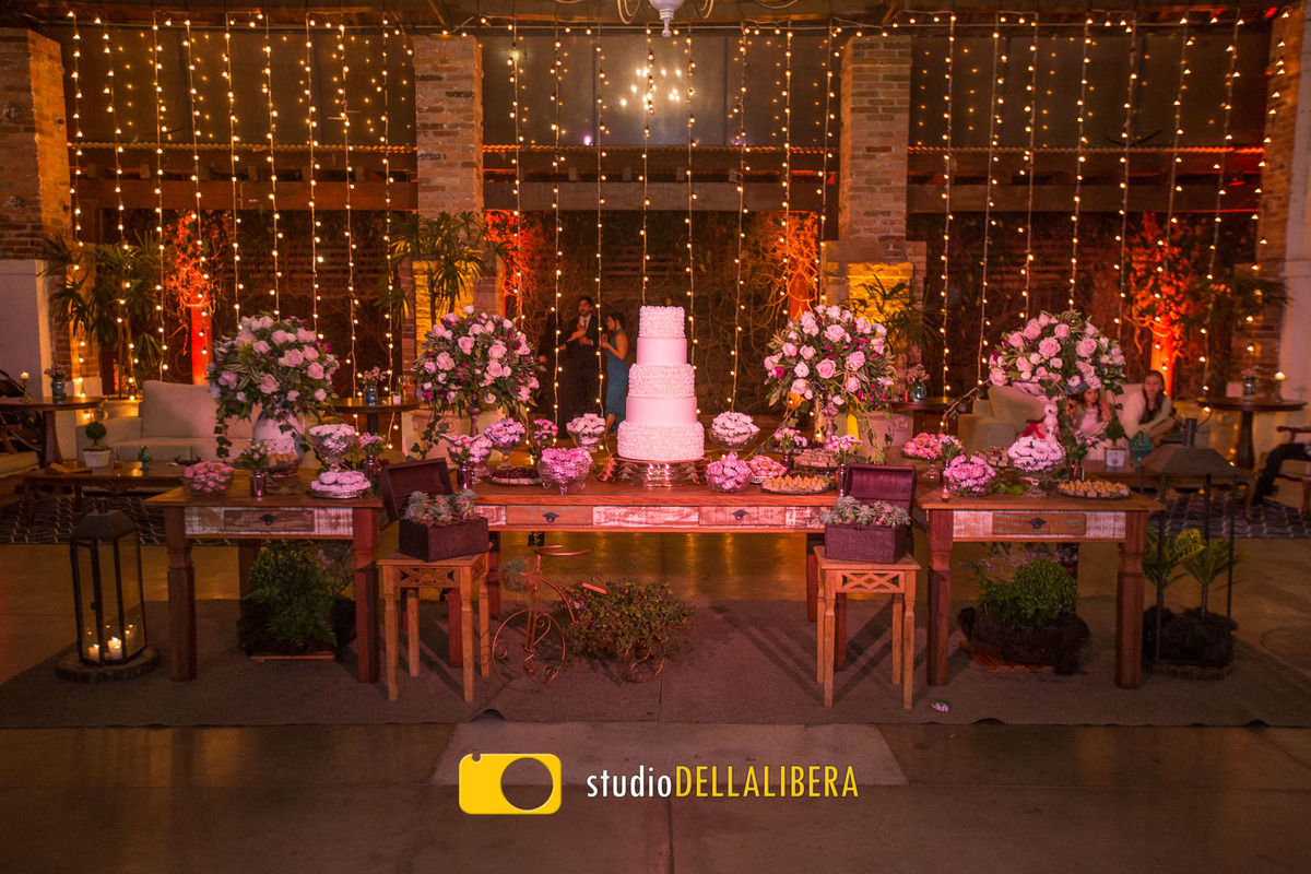 Foto da mesa do bolo do casamento de Ana e Rafa no espaço Zara em Piracicaba, decorado por Verde Total Eventos com flores brancas, Livros e bolo artístico ao fundo cortina de luz