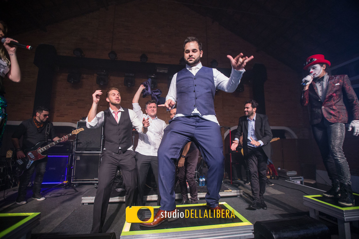 Noivo dançando em cima do palco da banda Viva Noite, com o cantor da banda ao fundo e os padrinhos atráz do noivo dançando 