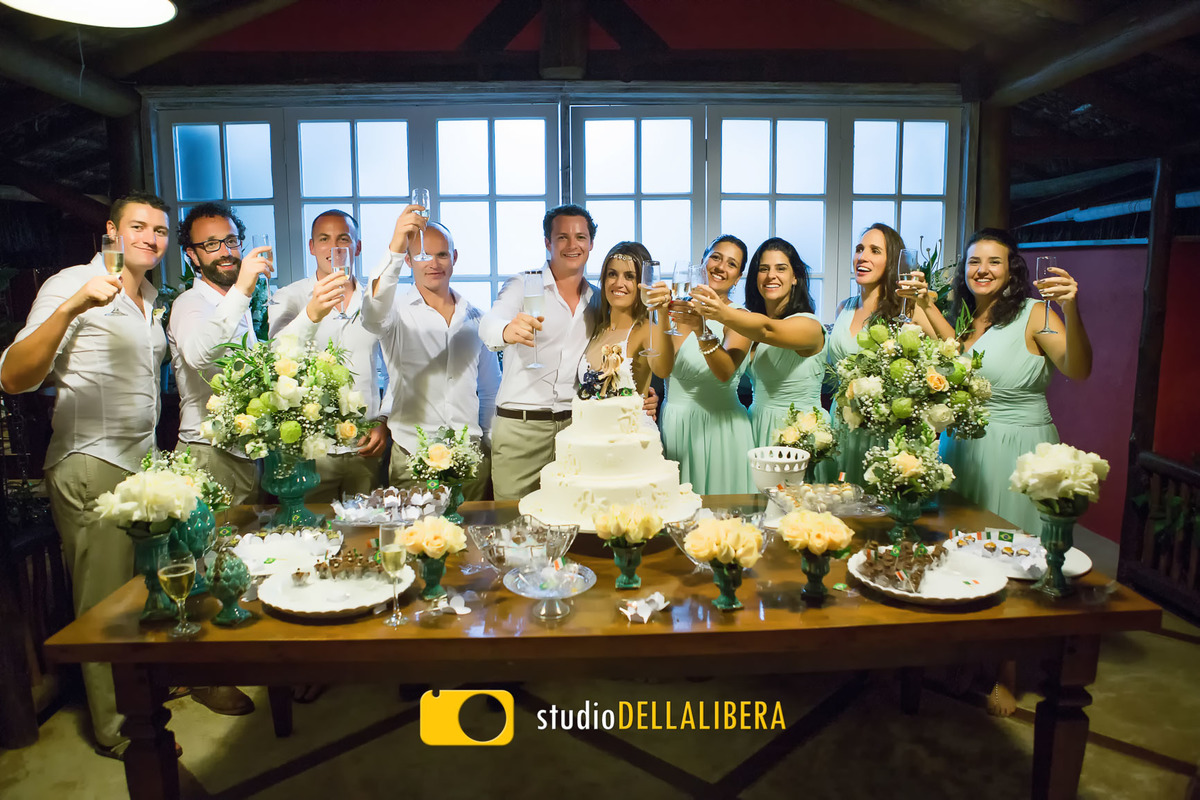 Mesa do bolo com os padrinhos e noivos brindando atrás da mesa que estava decorada com flores brancas, no casamento que foi realizado em Ilha Bela