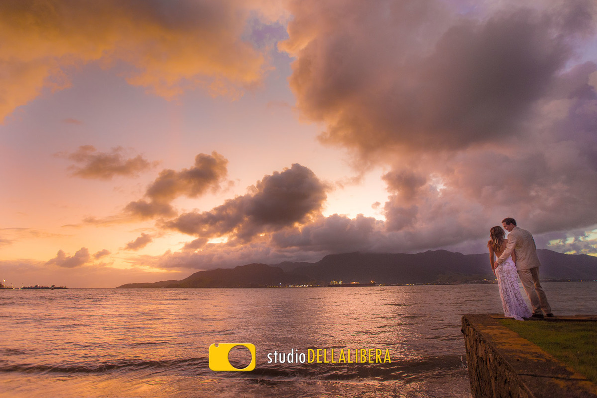 Noivos abraçados de costas admirando o mar de Ilha Bela na praia logo após o casamento com por do céu entre as nuvens 