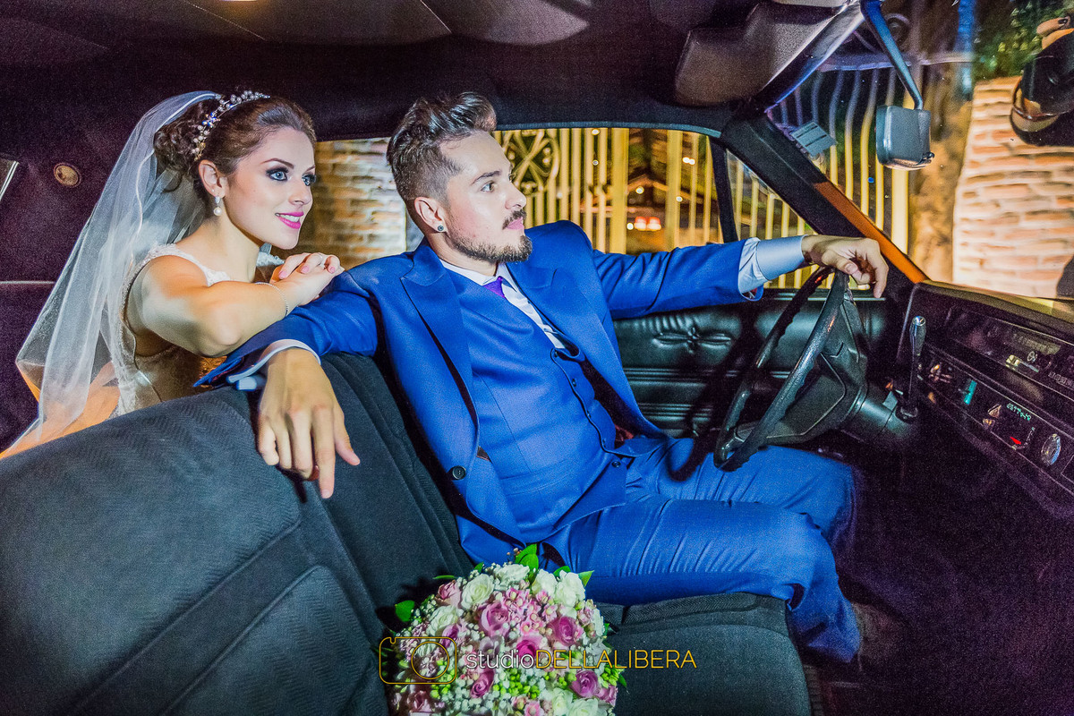 Noivo no volante de um carro com noiva de passageiro atrás dele e buque dela ao lado dele