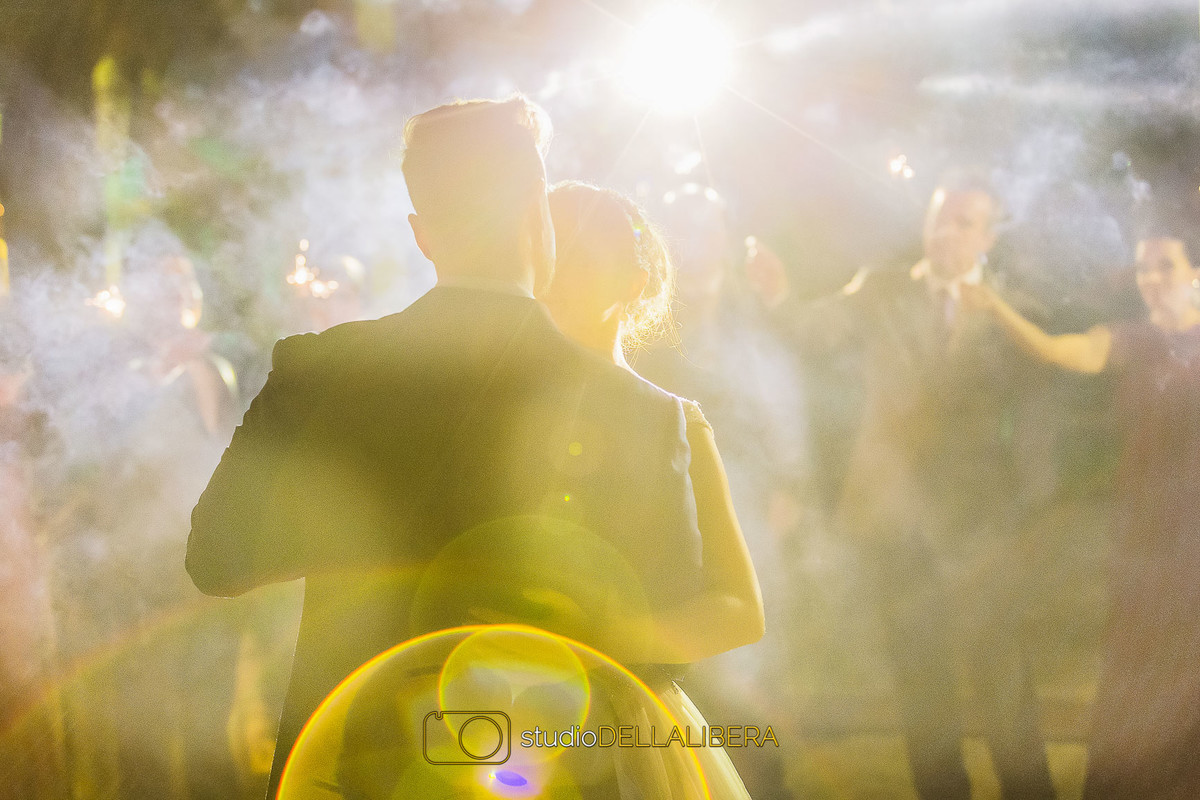 Noivo de costas beijando a testa da noiva e convidados dançando atrás