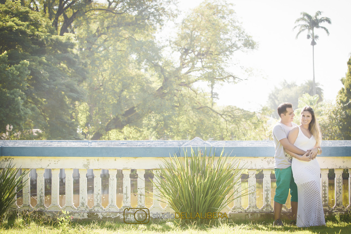 Noivo segurando a noiva por trás encostado na mureta com um lago como paisagem