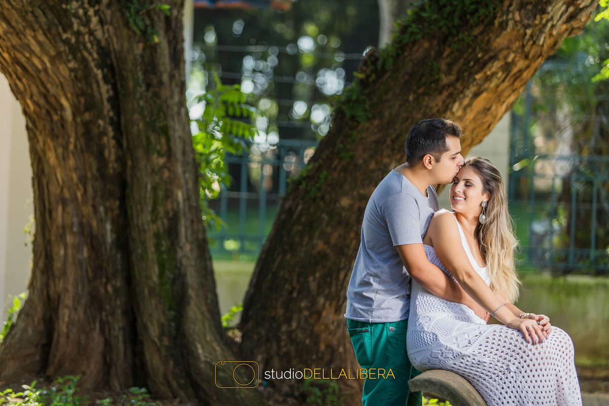 Noiva sentada num banco e o noivo com seus braços em sua cintura a beijando na testa