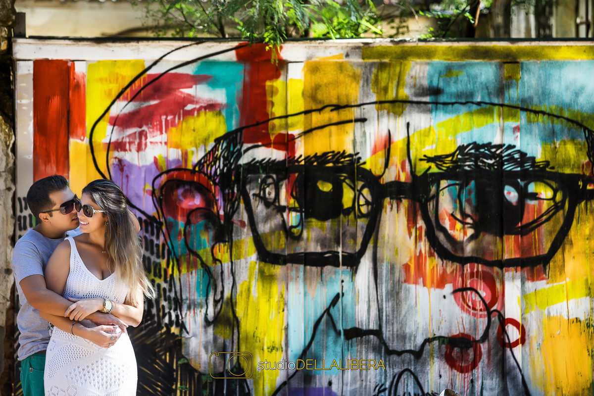 Noivo abraçando a noiva por trás tentado beijar o rosto dela encostados numa parede com desenho