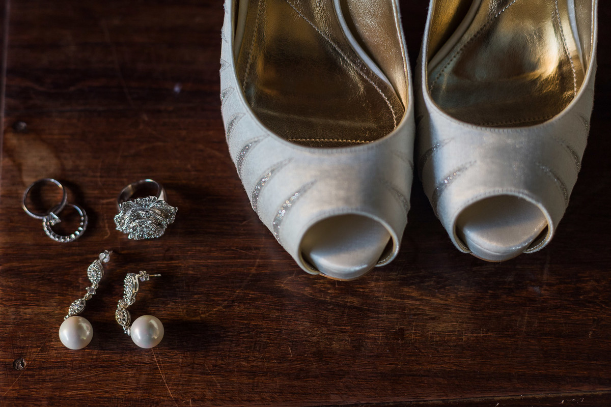 Sapatos da noiva junto com um brinco com pedrarias em prata e pérolas combinando com um anel em formato de rosa também com pedraria e um anel interligado com o outro em prata em com pedras