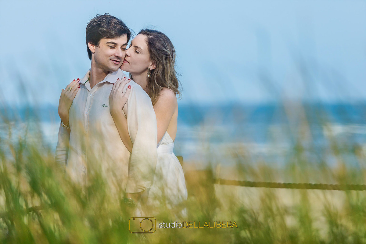 Noiva beijando e abraçando o noivo com plantas na frente e praia ao fundo, na sessão pre wedding na riviera de São Lourenço