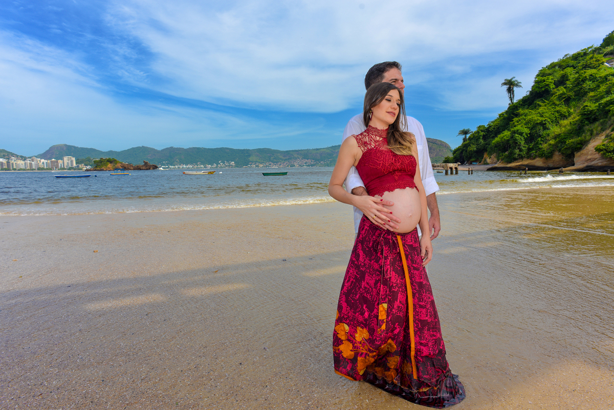 Ensaio fotográfico profissional gestante em niterói Nossa Casa Estúdio na praia de boa viagem no por do sol com saia longa e top vermelho mar calmo gestação gravidez mãe de primeira viagem com marido casal rio de janeiro primavera verão vestido maquiagem