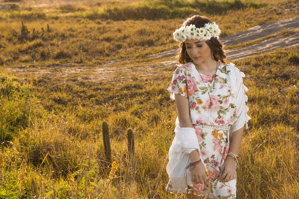 Ensaio fotográfico profissional de 15 anos na praia e no campo ao ar livre. Debutante de roupa branca e florida com tiara de flores na cabeça ao por do Sol, sorrindo. Festa Editorial Moda Revista Teen Adolescente Nossa Casa Estúdio Niterói Rio de Janeiro