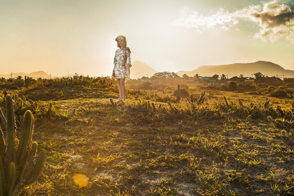 Ensaio fotográfico profissional de 15 anos na praia e no campo ao ar livre. Debutante de roupa branca e florida com tiara de flores na cabeça ao por do Sol, sorrindo. Festa Editorial Moda Revista Teen Adolescente Nossa Casa Estúdio Niterói Rio de Janeiro