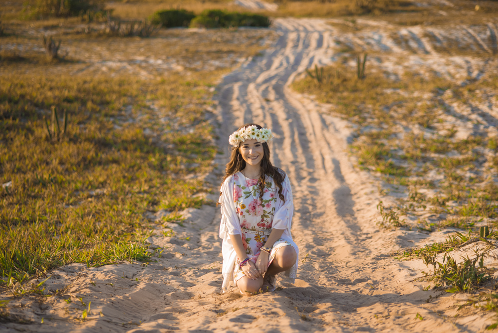 Ensaio fotográfico profissional de 15 anos na praia e no campo ao ar livre. Debutante de roupa branca e florida com tiara de flores na cabeça ao por do Sol, sorrindo. Festa Editorial Moda Revista Teen Adolescente Nossa Casa Estúdio Niterói Rio de Janeiro