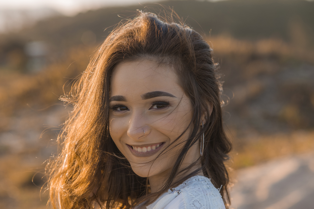 Ensaio fotográfico profissional de 15 anos na praia e no campo ao ar livre. Debutante de roupa branca e florida com tiara de flores na cabeça ao por do Sol, sorrindo. Festa Editorial Moda Revista Teen Adolescente Nossa Casa Estúdio Niterói Rio de Janeiro