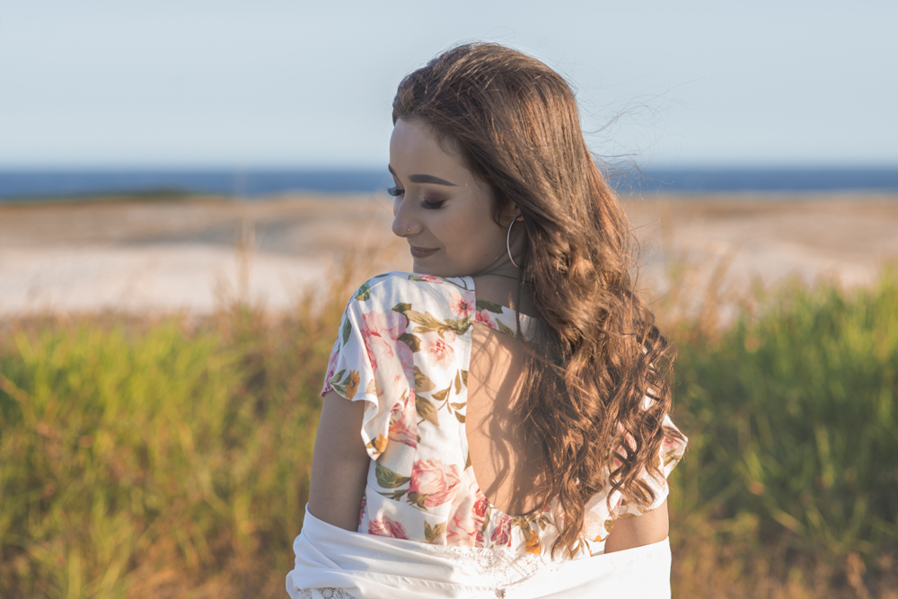 Ensaio fotográfico profissional de 15 anos na praia e no campo ao ar livre. Debutante de roupa branca e florida com tiara de flores na cabeça ao por do Sol, sorrindo. Festa Editorial Moda Revista Teen Adolescente Nossa Casa Estúdio Niterói Rio de Janeiro