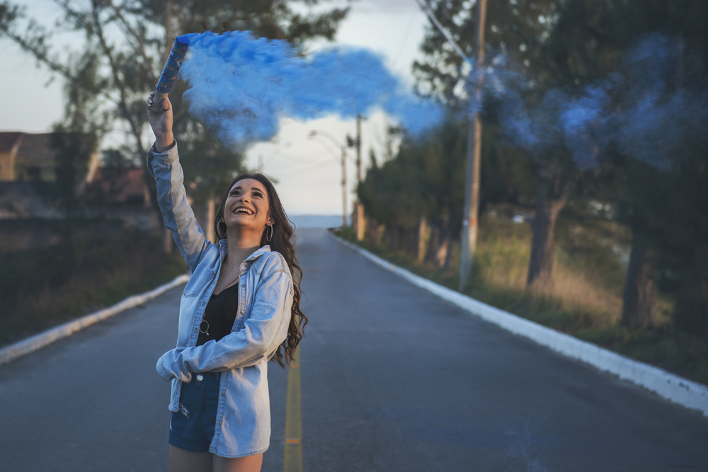 Ensaio fotográfico profissional de 15 anos fumaça e no campo ao ar livre. Debutante de roupa branca e florida com tiara de flores na cabeça ao por do Sol, sorrindo. Festa Editorial Moda Revista Teen Adolescente Nossa Casa Estúdio Niterói Rio de Janeiro