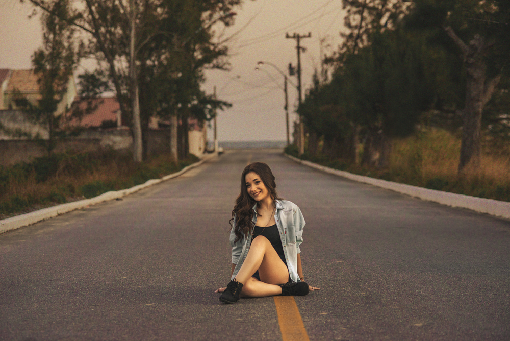 Ensaio fotográfico profissional de 15 anos na praia e no campo ao ar livre. Debutante de roupa branca e florida com tiara de flores na cabeça ao por do Sol, sorrindo. Festa Editorial Moda Revista Teen Adolescente Nossa Casa Estúdio Niterói Rio de Janeiro