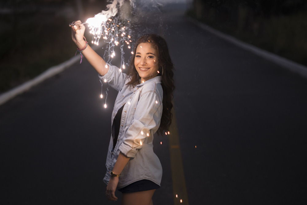 Ensaio fotográfico profissional de 15 anos na praia e faísca ao ar livre. Debutante de roupa branca e florida com tiara de flores na cabeça ao por do Sol, sorrindo. Festa Editorial Moda Revista Teen Adolescente Nossa Casa Estúdio Niterói Rio de Janeiro