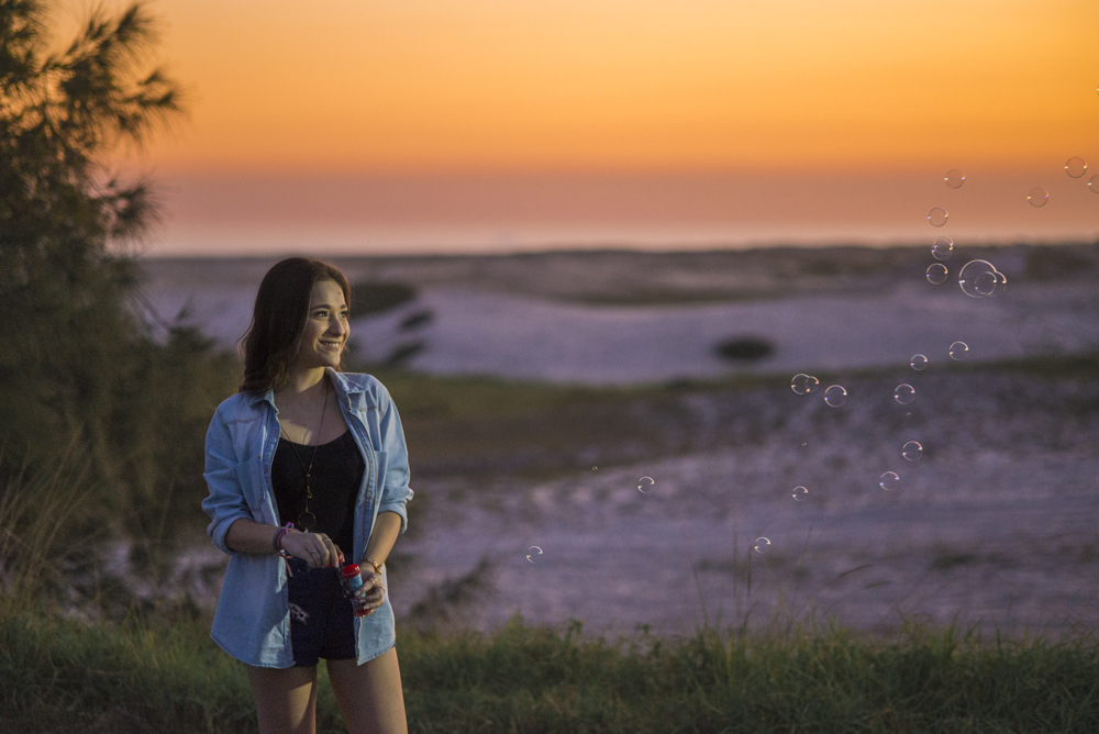 Ensaio fotográfico profissional de 15 anos na praia e no campo ao ar livre. Debutante de roupa branca e florida com tiara de flores na cabeça ao por do Sol, sorrindo. Festa Editorial Moda Revista Teen Adolescente Nossa Casa Estúdio Niterói Rio de Janeiro