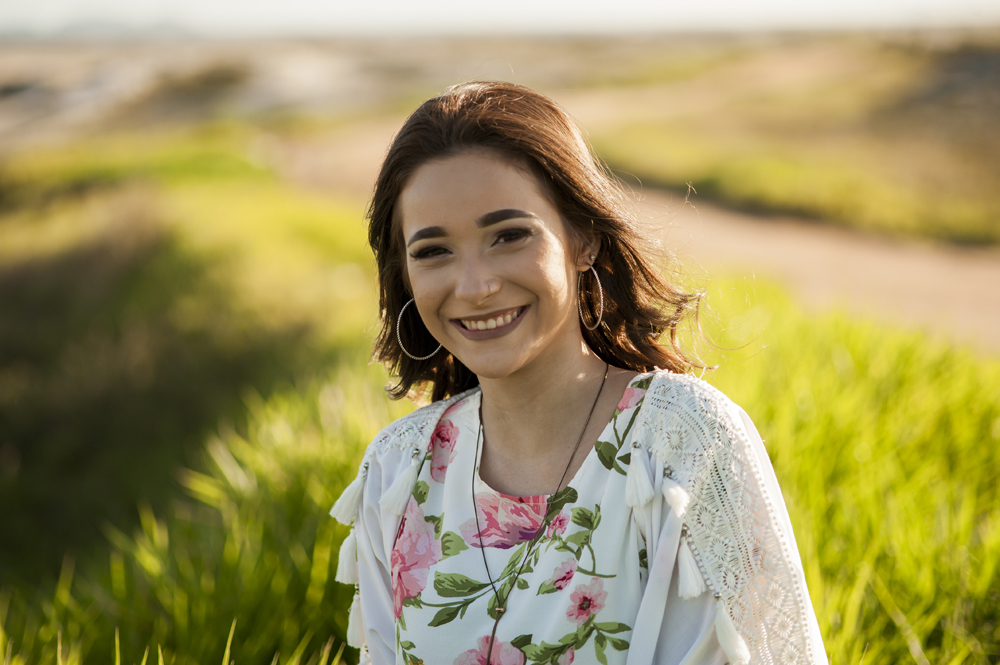 Ensaio fotográfico profissional de 15 anos na praia e no campo ao ar livre. Debutante de roupa branca e florida com tiara de flores na cabeça ao por do Sol, sorrindo. Festa Editorial Moda Revista Teen Adolescente Nossa Casa Estúdio Niterói Rio de Janeiro