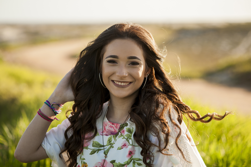 Ensaio fotográfico profissional de 15 anos na praia e no campo ao ar livre. Debutante de roupa branca e florida com tiara de flores na cabeça ao por do Sol, sorrindo. Festa Editorial Moda Revista Teen Adolescente Nossa Casa Estúdio Niterói Rio de Janeiro