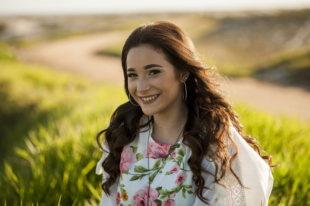 Ensaio fotográfico profissional de 15 anos na praia e no campo ao ar livre. Debutante de roupa branca e florida com tiara de flores na cabeça ao por do Sol, sorrindo. Festa Editorial Moda Revista Teen Adolescente Nossa Casa Estúdio Niterói Rio de Janeiro