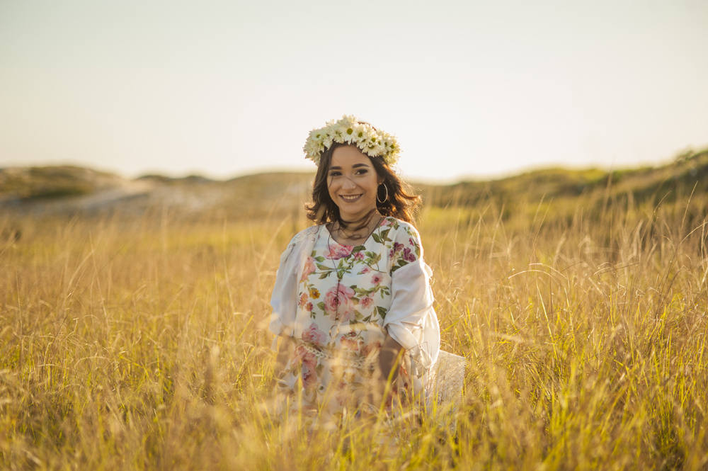 Ensaio fotográfico profissional de 15 anos na praia e no campo ao ar livre. Debutante de roupa branca e florida com tiara de flores na cabeça ao por do Sol, sorrindo. Festa Editorial Moda Revista Teen Adolescente Nossa Casa Estúdio Niterói Rio de Janeiro