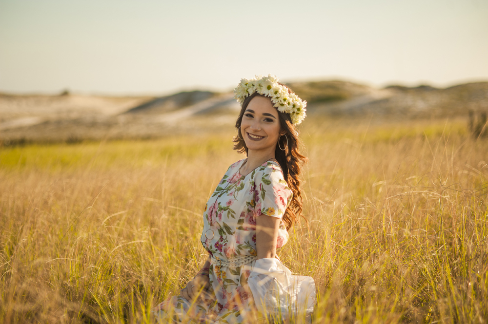 Ensaio fotográfico profissional de 15 anos na praia e no campo ao ar livre. Debutante de roupa branca e florida com tiara de flores na cabeça ao por do Sol, sorrindo. Festa Editorial Moda Revista Teen Adolescente Nossa Casa Estúdio Niterói Rio de Janeiro