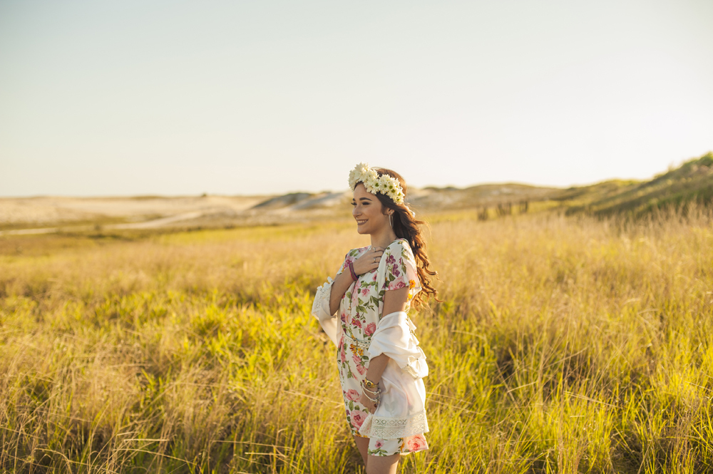 Ensaio fotográfico profissional de 15 anos na praia e no campo ao ar livre. Debutante de roupa branca e florida com tiara de flores na cabeça ao por do Sol, sorrindo. Festa Editorial Moda Revista Teen Adolescente Nossa Casa Estúdio Niterói Rio de Janeiro