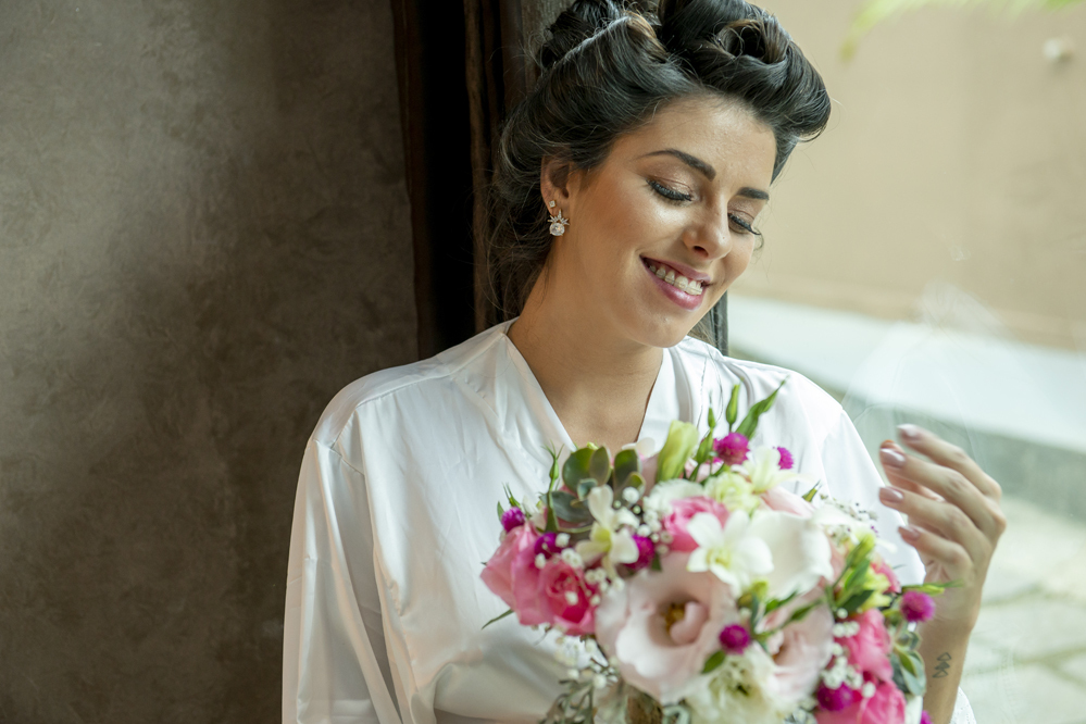 Fotográfo de casamento em Niterói Rio de Janeiro Maricá no recanto dos coqueiros noiva se maquiando com maquiadora em making of com vestido branco antes da cerimônia e festa com Nossa Casa Estúdio maquiagem da noiva batom cores no espelho Buquê de flores