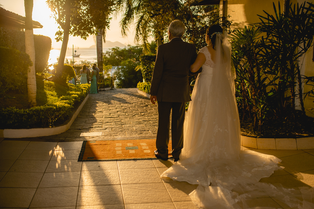 Nossa Casa Estúdio Fotografia de casamento em Niterói Rio de Janeiro Búzios Cabo Frio Hotel Atlântico Búzios noiva em cerimônia de casamento com pai antes da entrada com vestido branco no por do Sol de frente para  mar para a praia com decoração rústica