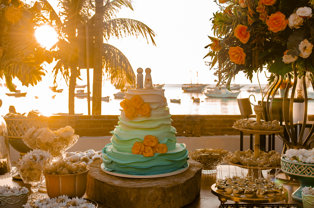 Nossa Casa Estúdio Fotografia de casamento profissional em Búzios Niterói Rio de Janeiro Decoração Rústica em festa no Hotel Atlântico Búzios Bolo de novo azul com flores laranja  com dicas e tendências a beira mar de dia no por do Sol laranja e docinhos