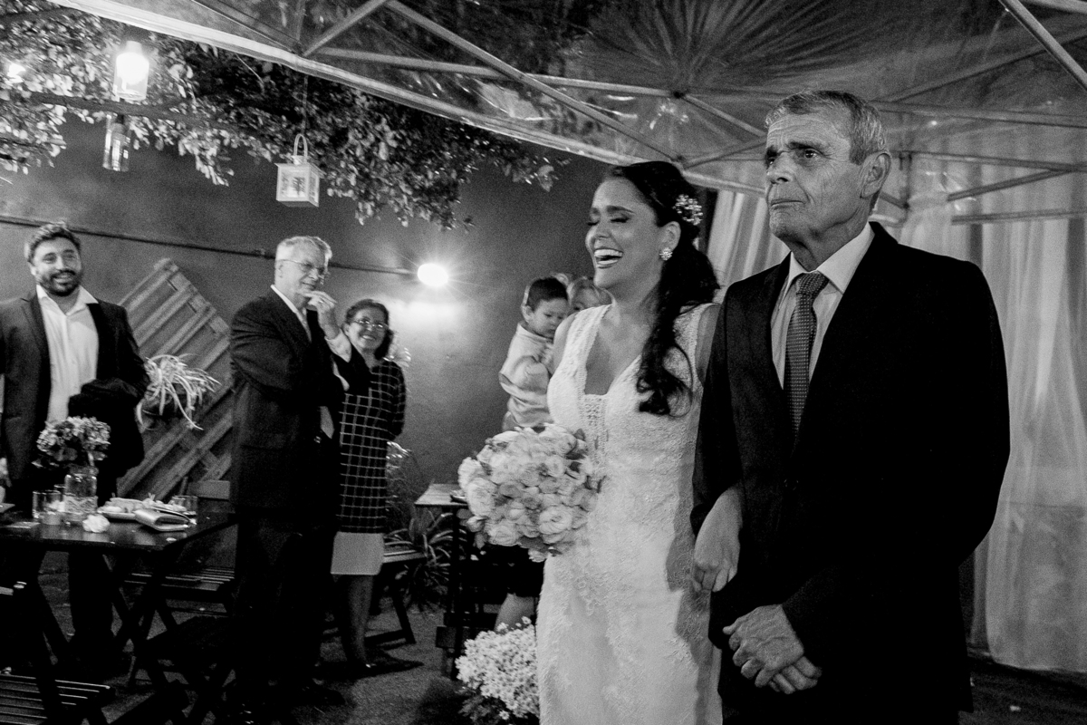 Nossa Casa estúdio fotografia de casamento em Niterói e no Rio de Janeiro entrada da noiva de vestido branco e maquiagem com pai de terno preto e convidados olhando com buquê de flores nas mãos fotografia preta e branca
