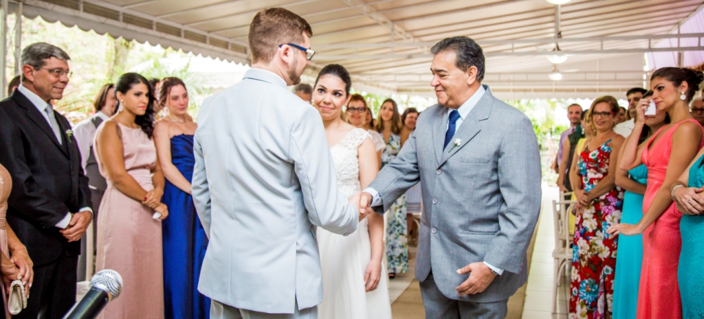 Pai da noiva com terno cinza apertando a mão do noivo com terno cinza azulado e noiva apaixonada olhando para o noivo. Cerimônia de casamento em Niterói
