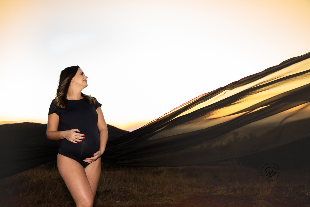 Gestante mamãe esperando bebe que irá nascer segurando barriga roupa preta body  por do sol tecido voando  por do sol