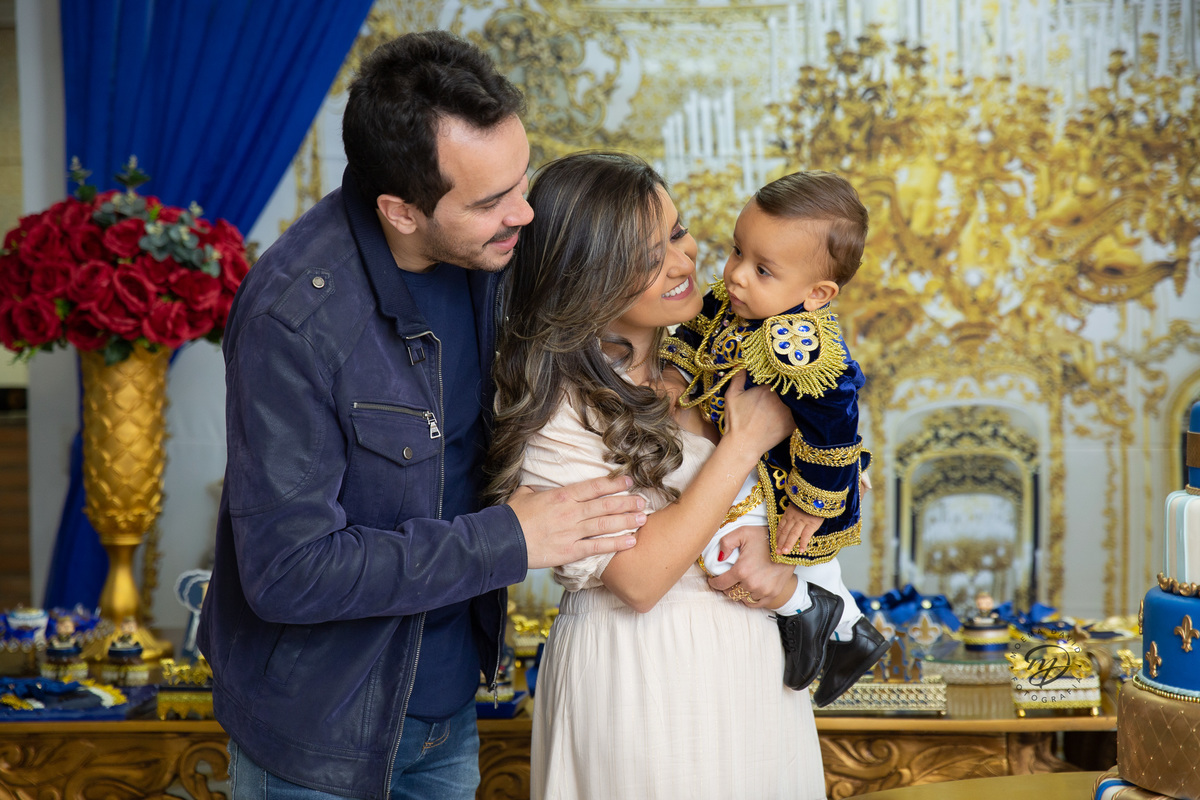 Príncipe Henrique no colo da mamãe e do papai com sua roupa de luxo olhando para seus pais