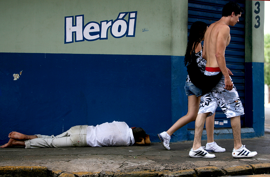 Morador de rua dorme no chão da calçada embaixo da palavra herói, permite interpretação semiótica